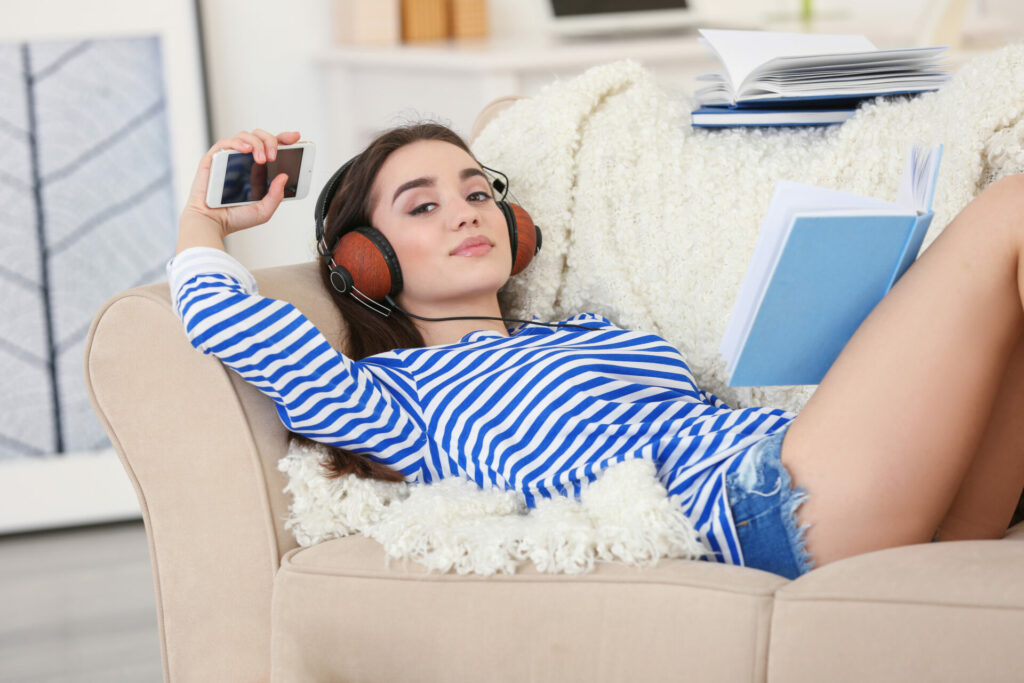 Eine junge Frau liegt entspannt auf einem Sofa, trägt Kopfhörer und hält ein Smartphone in der Hand. Neben ihr liegt ein aufgeschlagenes Buch, das sie offensichtlich liest.