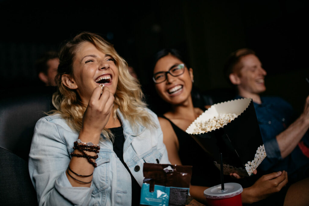 Eine Gruppe von Freunden sitzt in einem Kino und lacht fröhlich, während sie einen Film ansehen. Die Person im Vordergrund isst Popcorn und hat einen Softdrink neben sich.