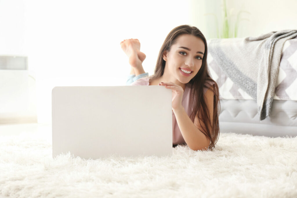 Eine junge Frau liegt auf einem weichen, flauschigen Teppich in einem gemütlichen Raum und lächelt in die Kamera. Vor ihr steht ein Laptop, auf den sie sich stützt. Im Hintergrund ist ein Bett mit einer gefalteten Decke zu sehen.