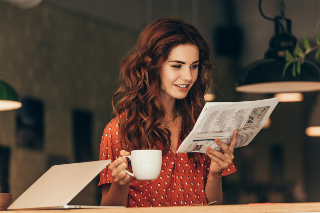 Eine junge Frau mit lockigem roten Haar sitzt in einem Café und liest eine Zeitung, während sie eine Tasse Kaffee in der Hand hält. Vor ihr steht ein aufgeklappter Laptop.