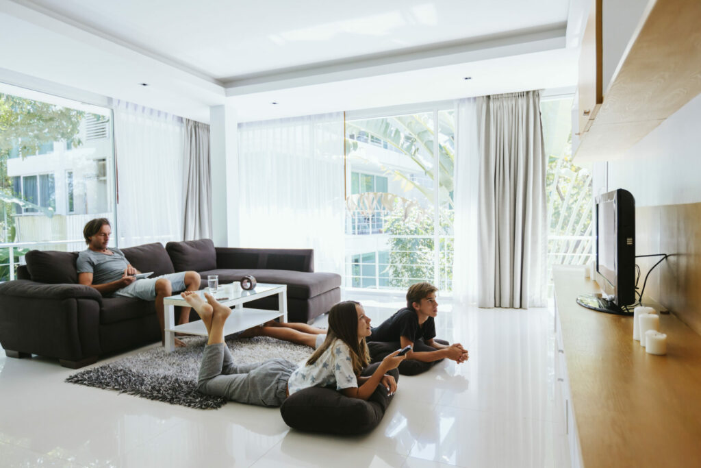 Eine Familie entspannt sich in einem modernen Wohnzimmer mit großen Fenstern. Zwei Kinder liegen auf dem Boden vor einem Fernseher, während der Vater auf dem Sofa sitzt und ein Tablet benutzt.