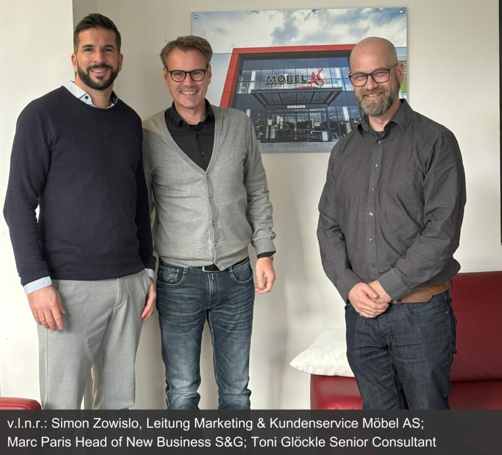 Unsere Mitarbeiter Marc Paris und Toni Glöckle stehen zusammen mit Simon Zowislo, Leitung Marketing & Kundenservice Möbel AS zusammen vor einer Wand auf dem ein Bild des Möbelhauses ist.